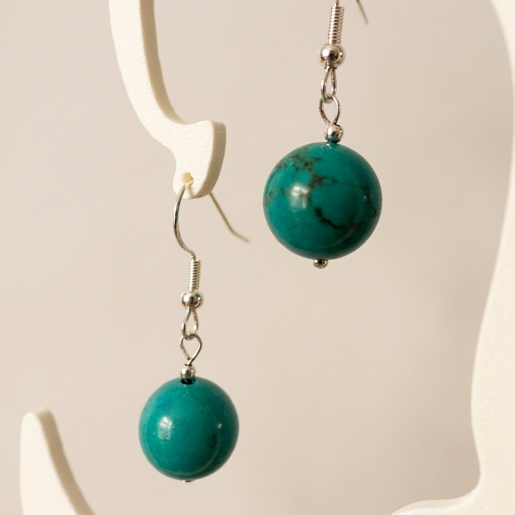Close-up of stabilised Hubei turquoise drop earrings hanging from a cream display stand, showing dark stone markings and sterling silver hooks.