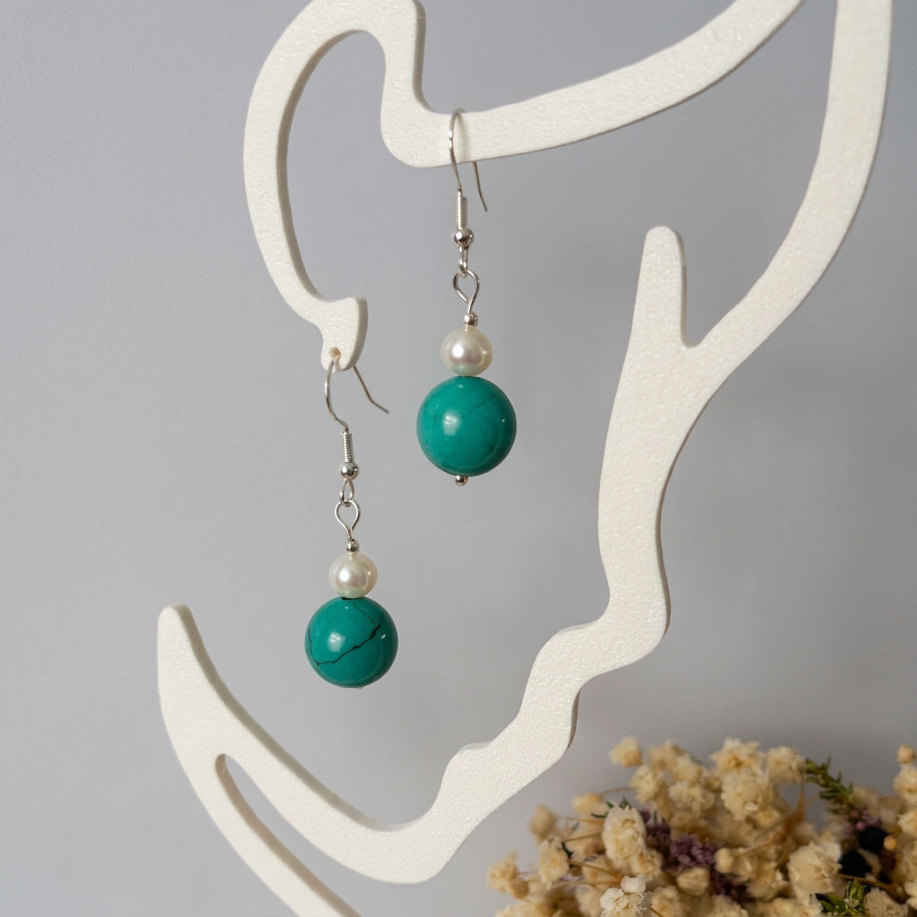 White baroque pearl and Hubei turquoise earrings hanging from a cream display stand beside dried flowers.