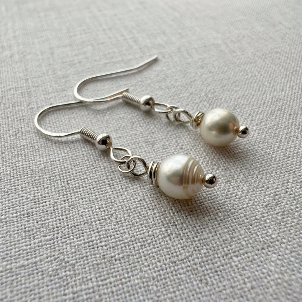 Close-up of white baroque pearl drop earrings on pale linen, showing a ridged pearl surface, sterling silver hooks, connecting rings and small silver bead tips.