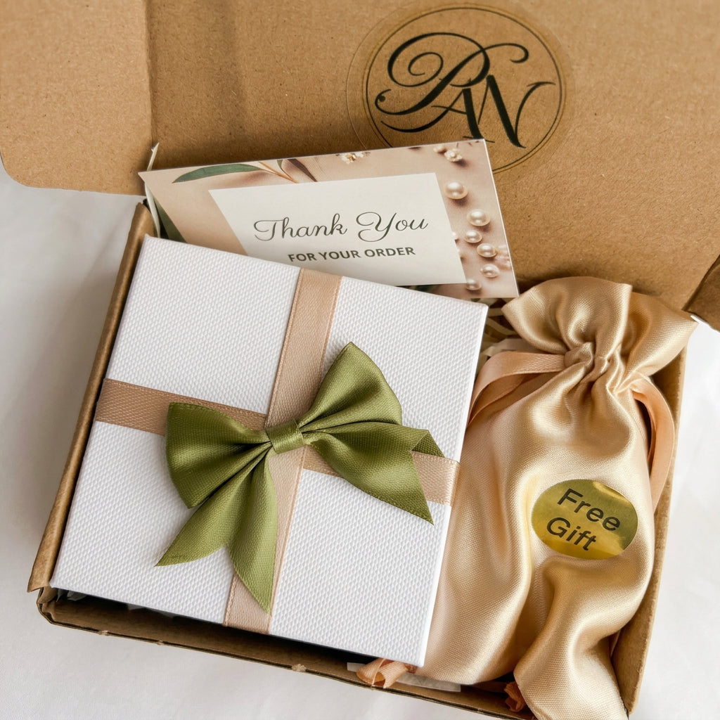 White jewellery gift box with beige ribbon and a green satin bow, thank-you card and gold satin pouch labelled “Free Gift”, inside a kraft postal box.