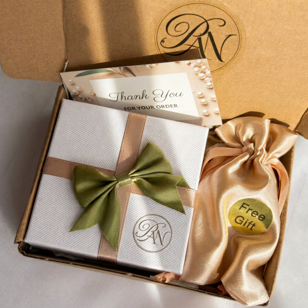 White jewellery gift box with beige ribbon and a green satin bow, thank-you card and gold satin pouch labelled “Free Gift”, inside a kraft postal box.