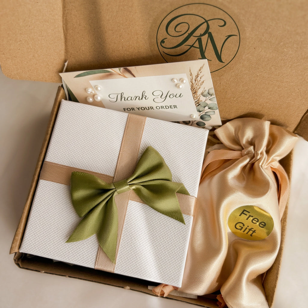 White jewellery gift box with beige ribbon and a green satin bow, thank-you card and gold satin pouch labelled “Free Gift”, inside a kraft postal box.