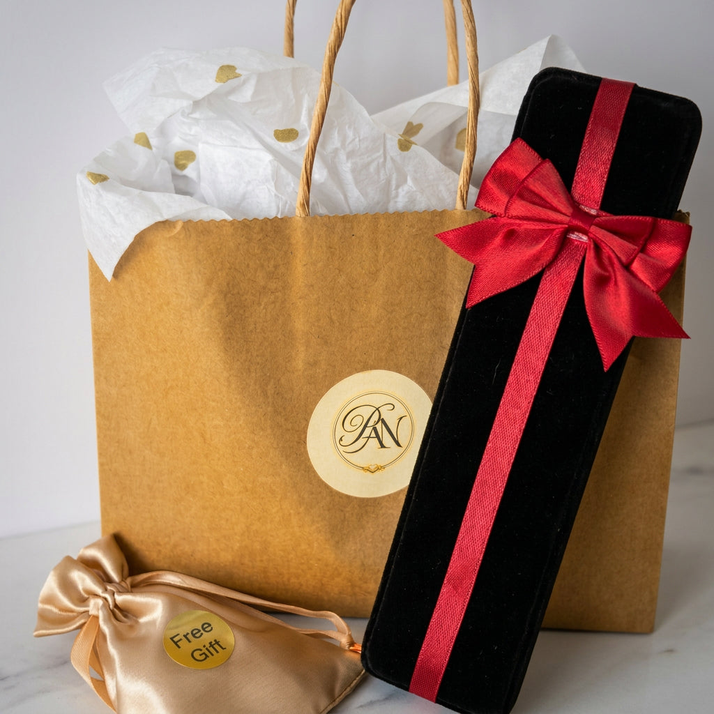 Gift packaging with a black velvet jewellery box tied with a red satin bow, a branded kraft paper bag filled with gold-heart tissue paper, and a champagne-coloured satin pouch labelled “Free Gift”.