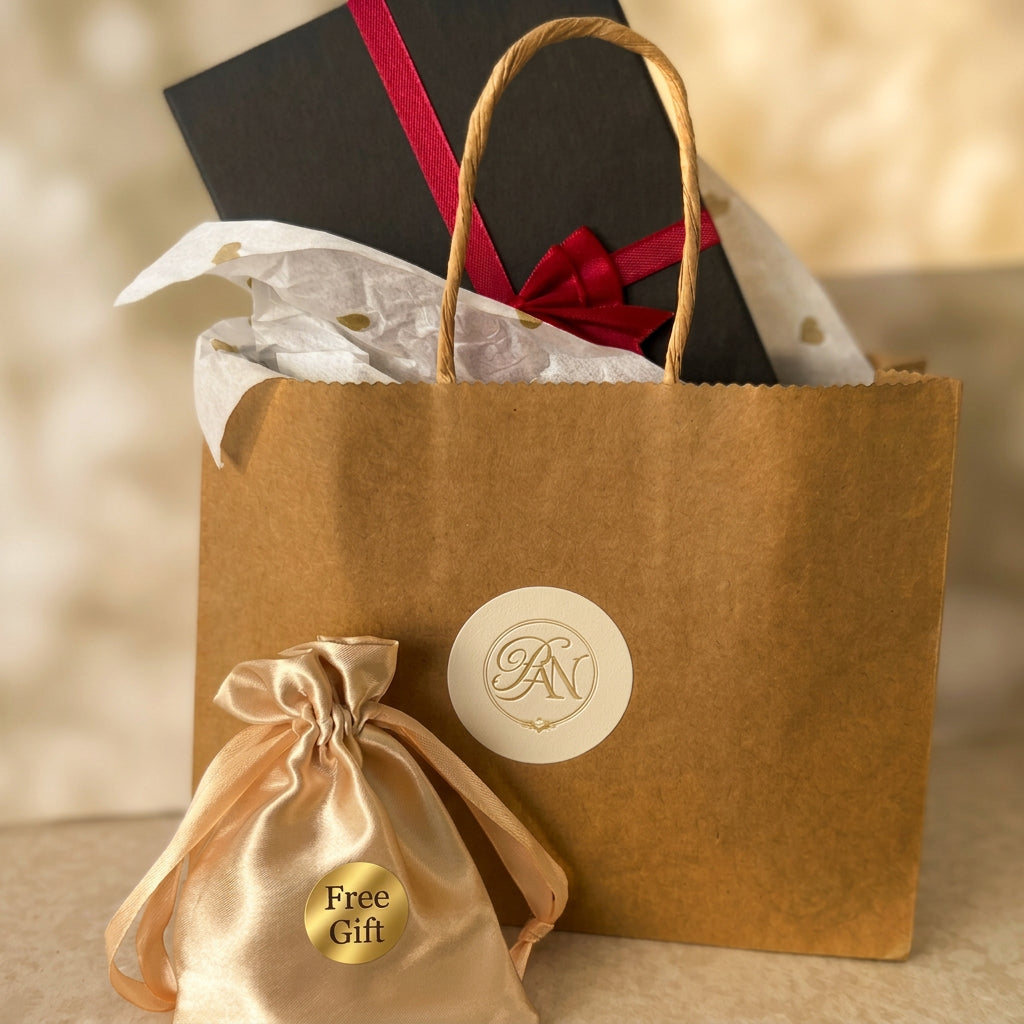 Branded kraft gift bag with white tissue paper and a black jewellery box tied with red ribbon, beside a gold satin pouch labelled “Free Gift”.