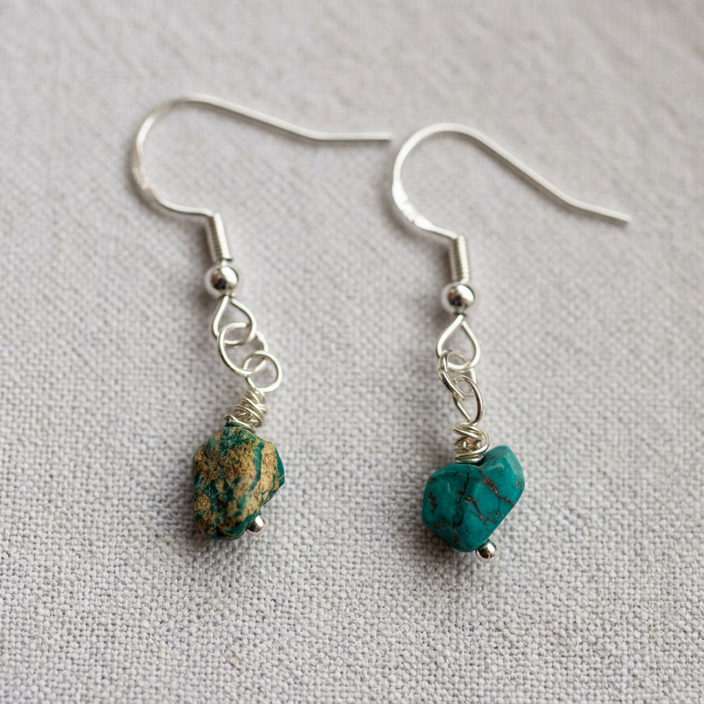 Raw Hubei turquoise drop earrings on cream linen, showing contrasting blue-green stones with brown matrix, wire wrapping and sterling silver hooks.