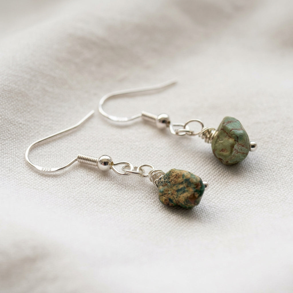 Pair of raw Hubei turquoise drop earrings with irregular green stones, brown matrix and sterling silver hooks, arranged on cream fabric.