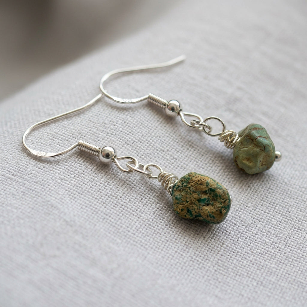 Raw Hubei turquoise drop earrings with irregular green-blue stones and earthy brown markings, wire-wrapped beneath sterling silver hooks on cream linen.⠋