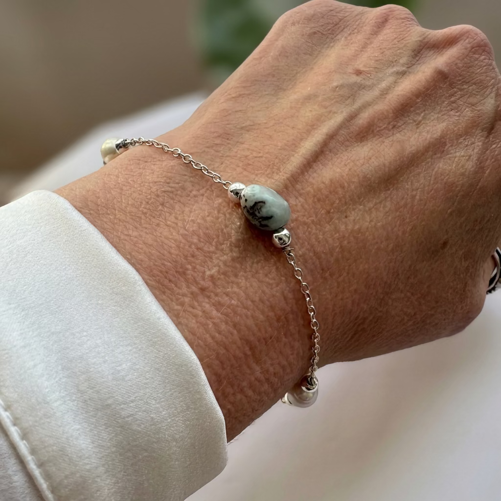 Delicate sterling silver chain bracelet with a pale blue larimar stone showing dark natural markings, polished silver beads and white baroque pearl accents, worn beside a white silk shirt cuff.