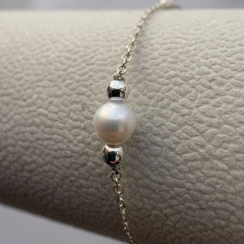 Close-up of a lustrous white baroque pearl between polished sterling silver spacers on a chain bracelet on a cream display.