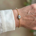 Delicate gold-filled chain bracelet with a pale blue larimar stone showing dark natural markings and white pearl accents, worn on the wrist beside a white silk shirt cuff.