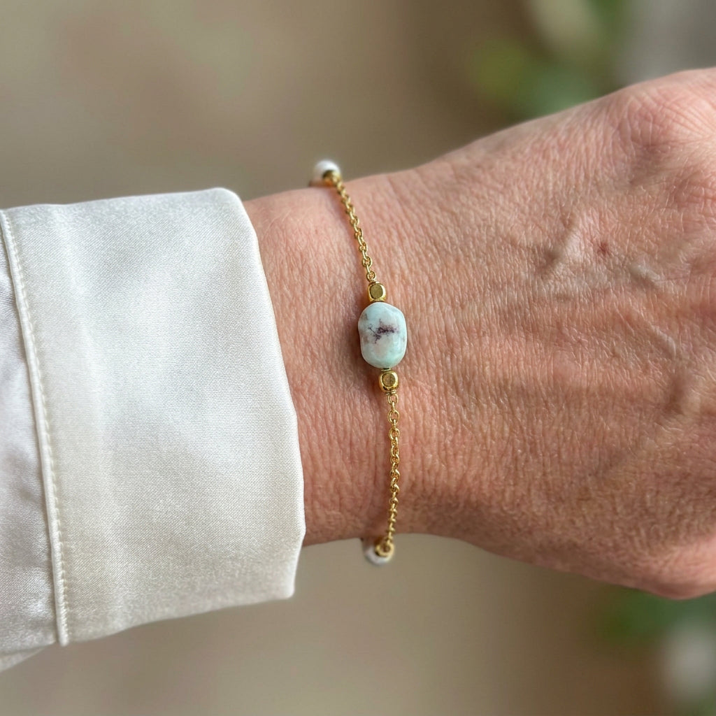 Delicate gold-filled chain bracelet with a pale blue larimar stone showing dark natural markings and white pearl accents, worn on the wrist beside a white silk shirt cuff.