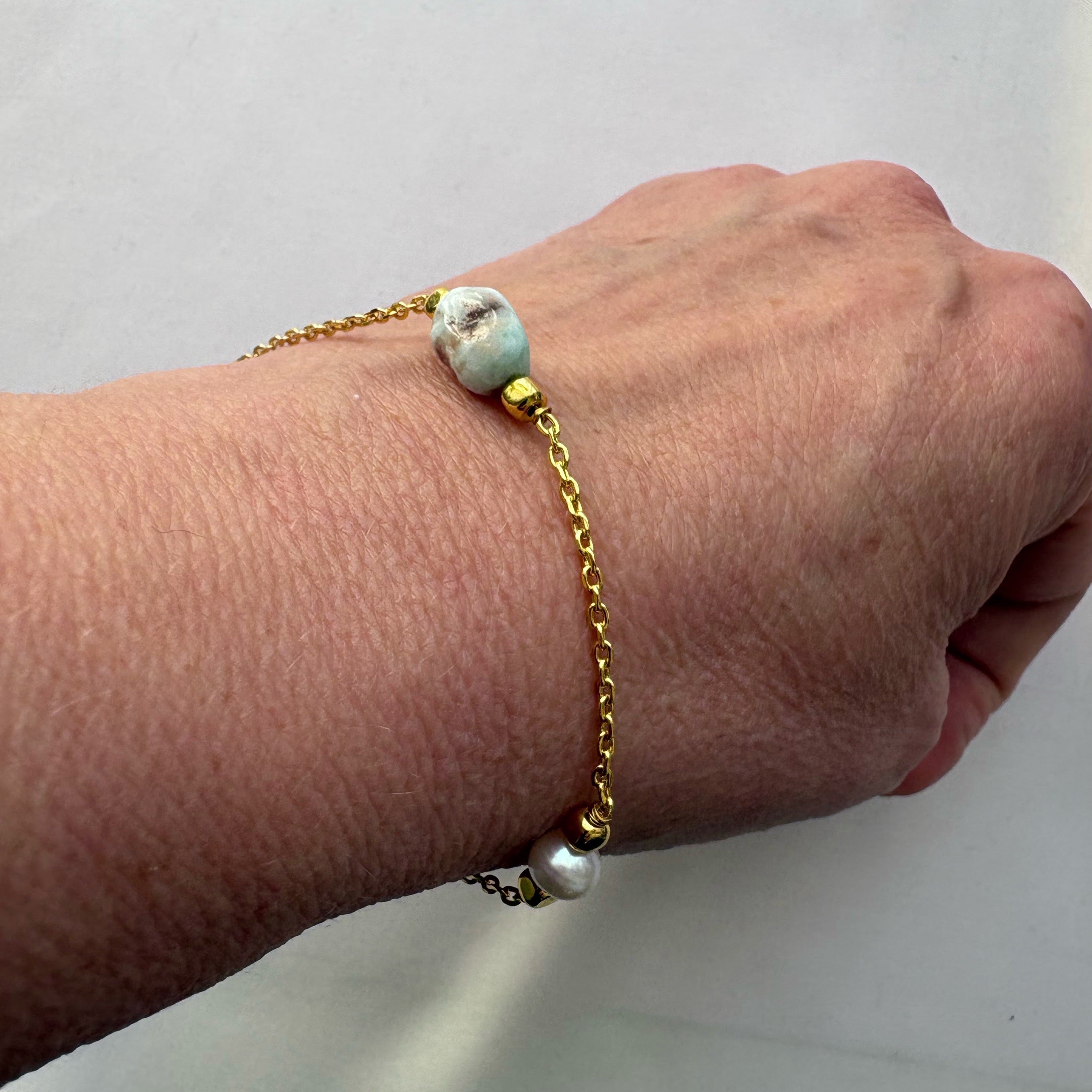 Gold-filled chain bracelet worn on a wrist, with a pale blue larimar bead above and a white baroque pearl below.