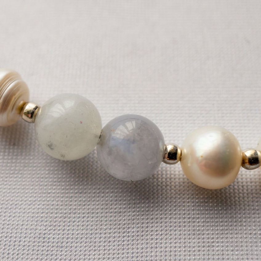 Macro view of pale aquamarine beads, a white baroque pearl and sterling silver spacers on cream fabric.