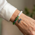 White and green jade bracelet with blue tourmaline beads, a snowflake charm and a colourful enamel advent window charm, worn on the wrist beside a white silk shirt cuff.