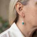 White baroque pearl and turquoise drop earring worn on the ear, with a small white pearl above a larger green-blue turquoise bead and sterling silver hook.