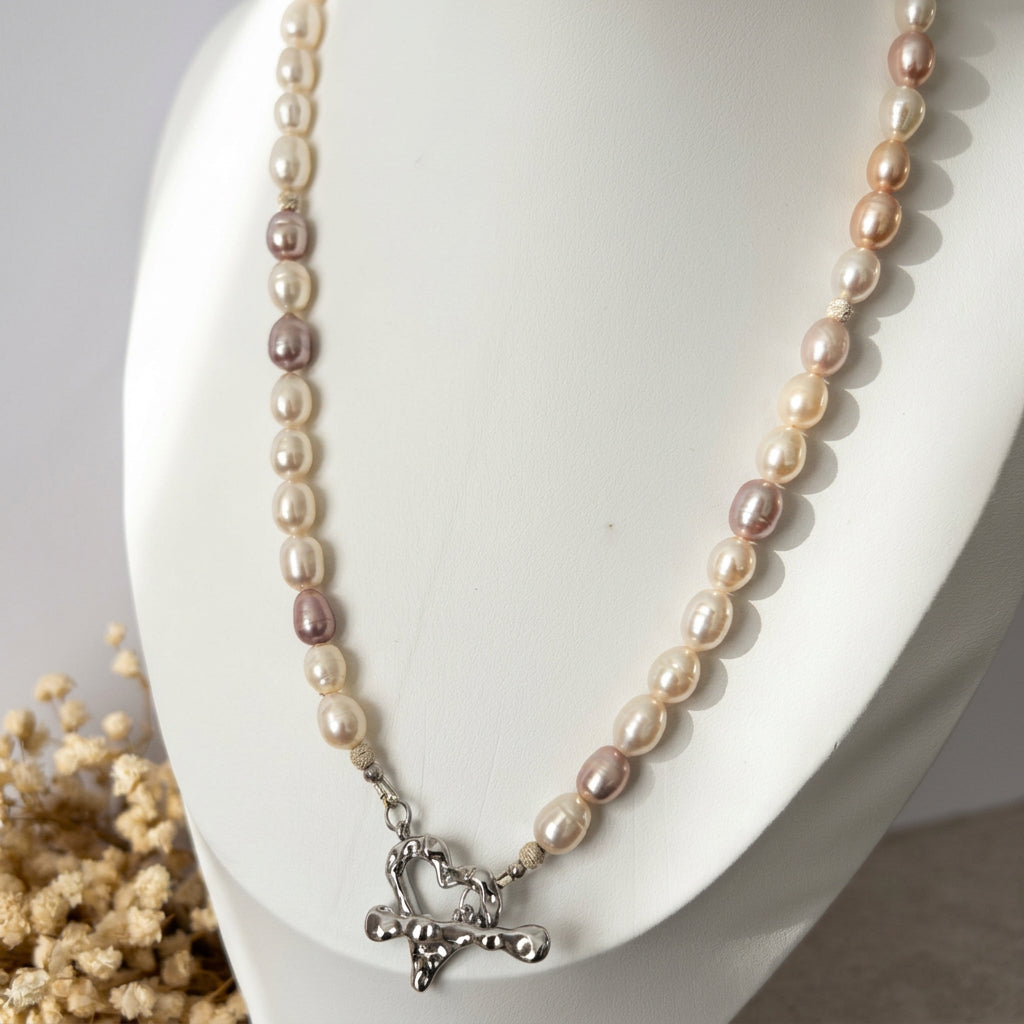 Creamy white and mauve-toned rice pearl necklace displayed on a white bust beside dried flowers, with small sterling silver accents and a fastened stainless steel heart-shaped toggle clasp at the front.