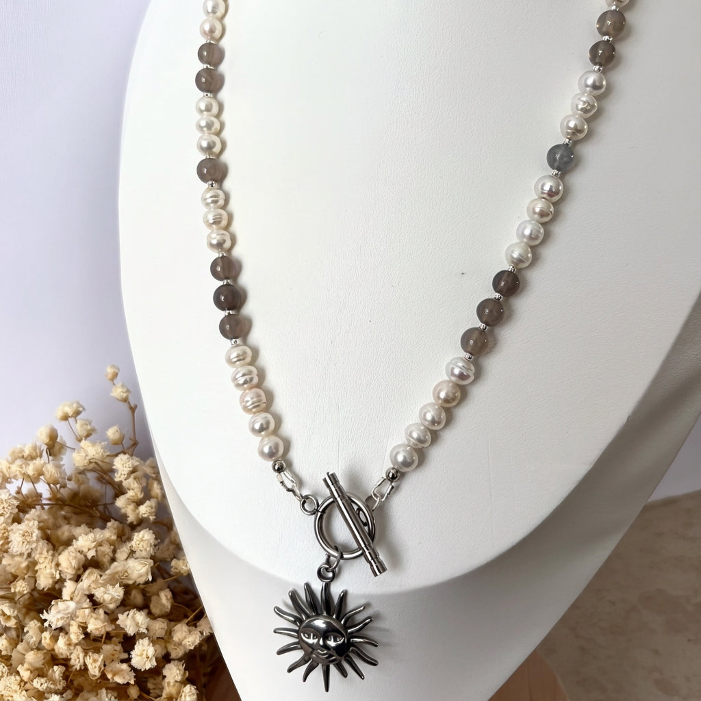 White baroque pearl and grey labradorite necklace displayed on a white bust beside dried cream flowers, featuring a fastened front toggle clasp and a stainless steel sun face pendant.