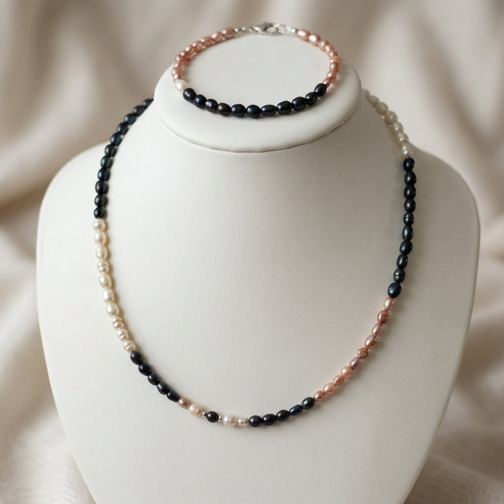 Matching rice pearl necklace and bracelet displayed on a white jewellery bust, with distinct sections of blush-pink, creamy white and dark blue pearls against a softly draped cream background.