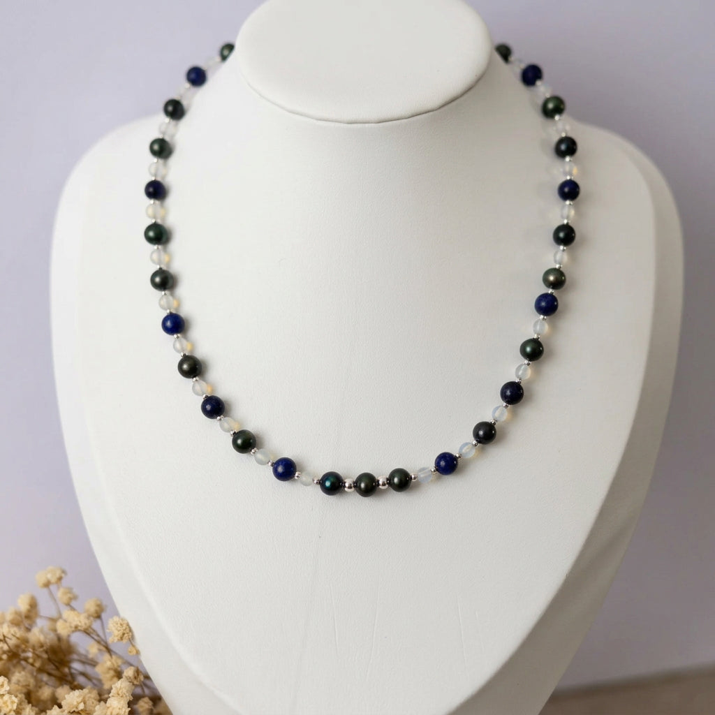 Peacock pearl, lapis lazuli and moonstone necklace displayed on a white bust beside dried cream flowers, showing dark green pearls, deep blue stones, pale translucent beads and small sterling silver spacers.⠋