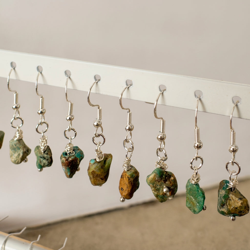 Close-up of raw Hubei turquoise drop earrings on a white rack, showing varied green and pale blue stones with brown matrix, silver wire wrapping and sterling silver hooks.