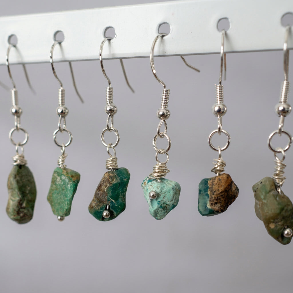 Close-up of raw Hubei turquoise drop earrings on a white rack, showing varied green and pale blue stones with brown matrix, silver wire wrapping and sterling silver hooks.