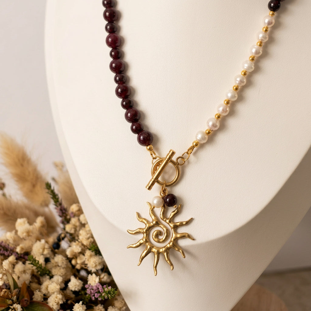 Burgundy garnet and white baroque pearl necklace with a gold-plated front toggle clasp, pearl and garnet drops, and a gold-coloured spiral sun pendant, displayed on a white jewellery bust beside dried flowers.