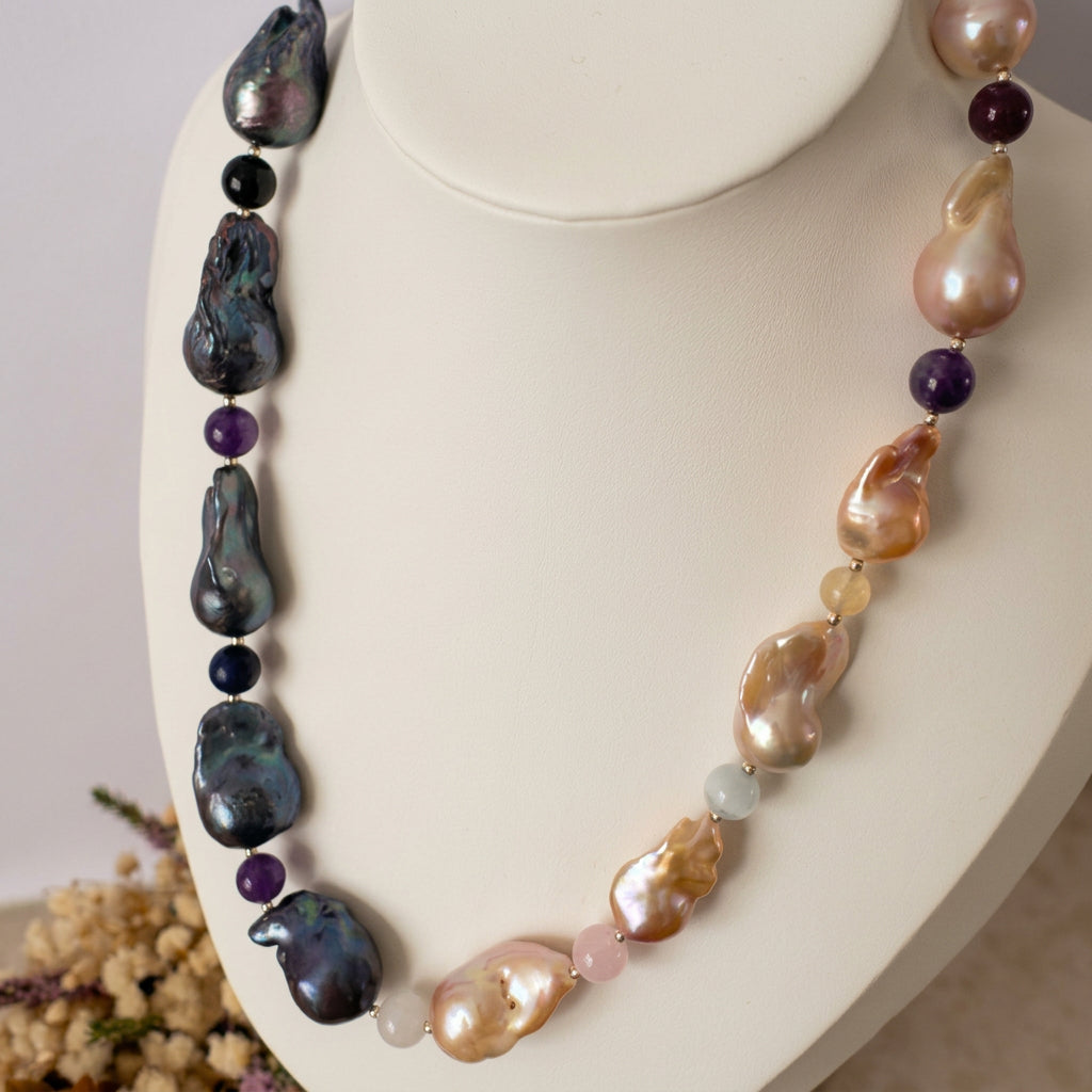 Statement necklace with dark blue-grey keshi pearls on one side and golden-cream keshi pearls on the other, separated by multicoloured gemstone beads, displayed on a white jewellery bust beside dried flowers.