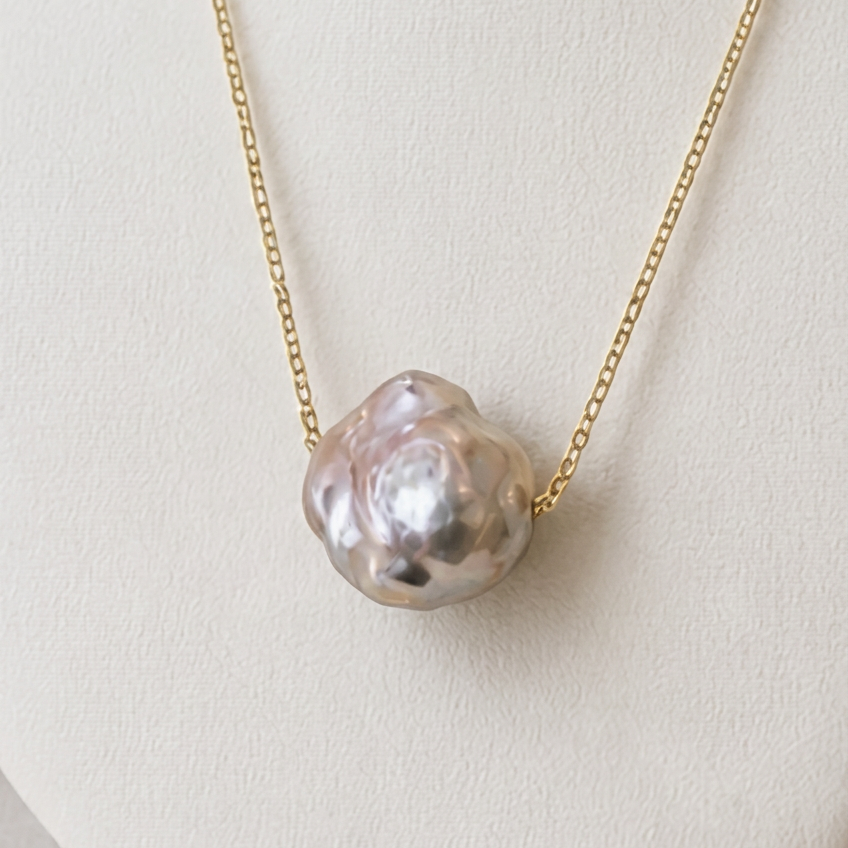 Close-up of a silvery-blush keshi pearl on a fine gold-over-sterling-silver chain against a white jewellery display, showing its rounded, rippled surface and pink-silver lustre.