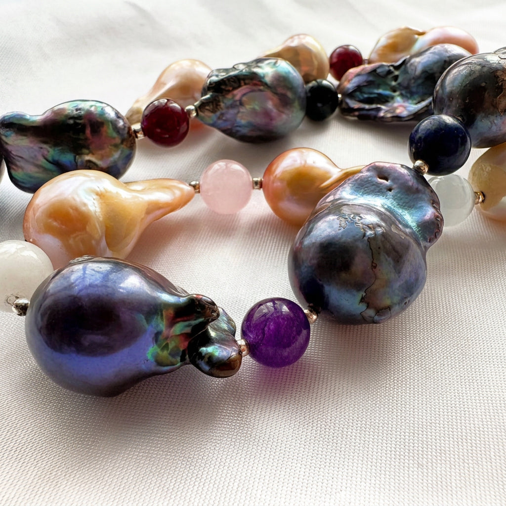 Close-up of large dark blue-grey and golden-cream keshi pearls with purple, blue, red and pale pink gemstone beads on cream fabric, showing the pearls’ irregular shapes and colourful iridescence.