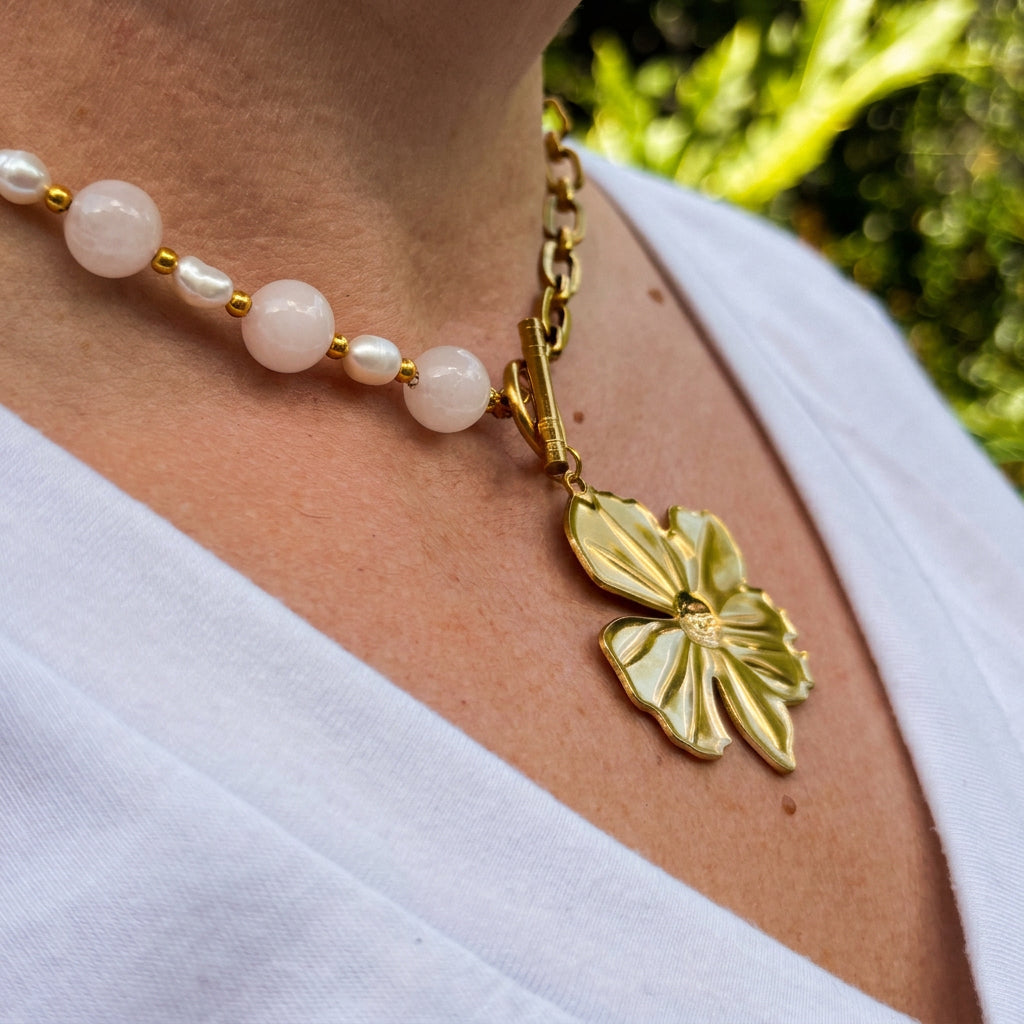 Angled close-up of pink quartz beads and white baroque pearls meeting a gold-plated paperclip chain at a front toggle clasp, with a large flower pendant worn over a white V-neck top.