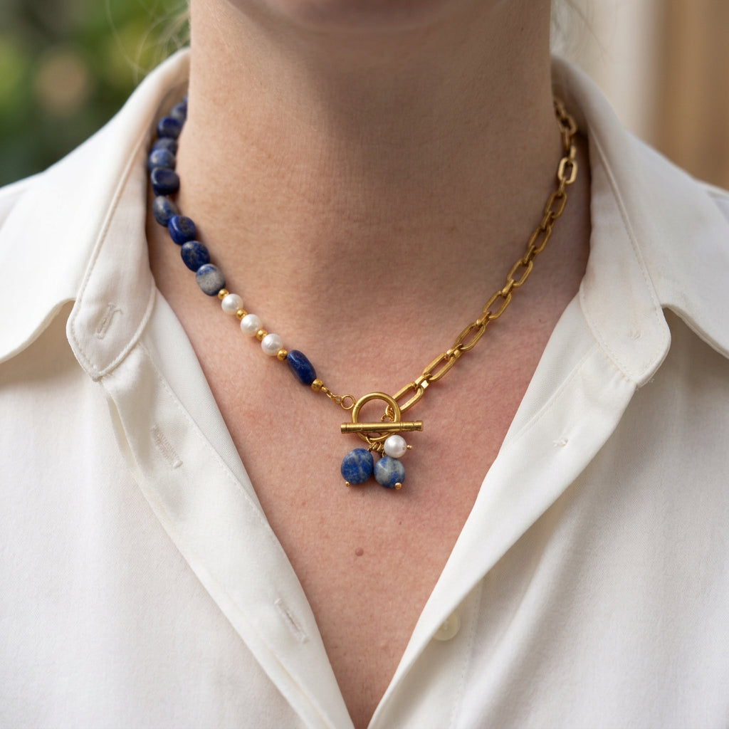 Blue lapis lazuli and white baroque pearl necklace with a gold-plated paperclip chain, fastened front toggle and a cluster of two lapis drops and one pearl, worn with an open-neck white shirt.⠋