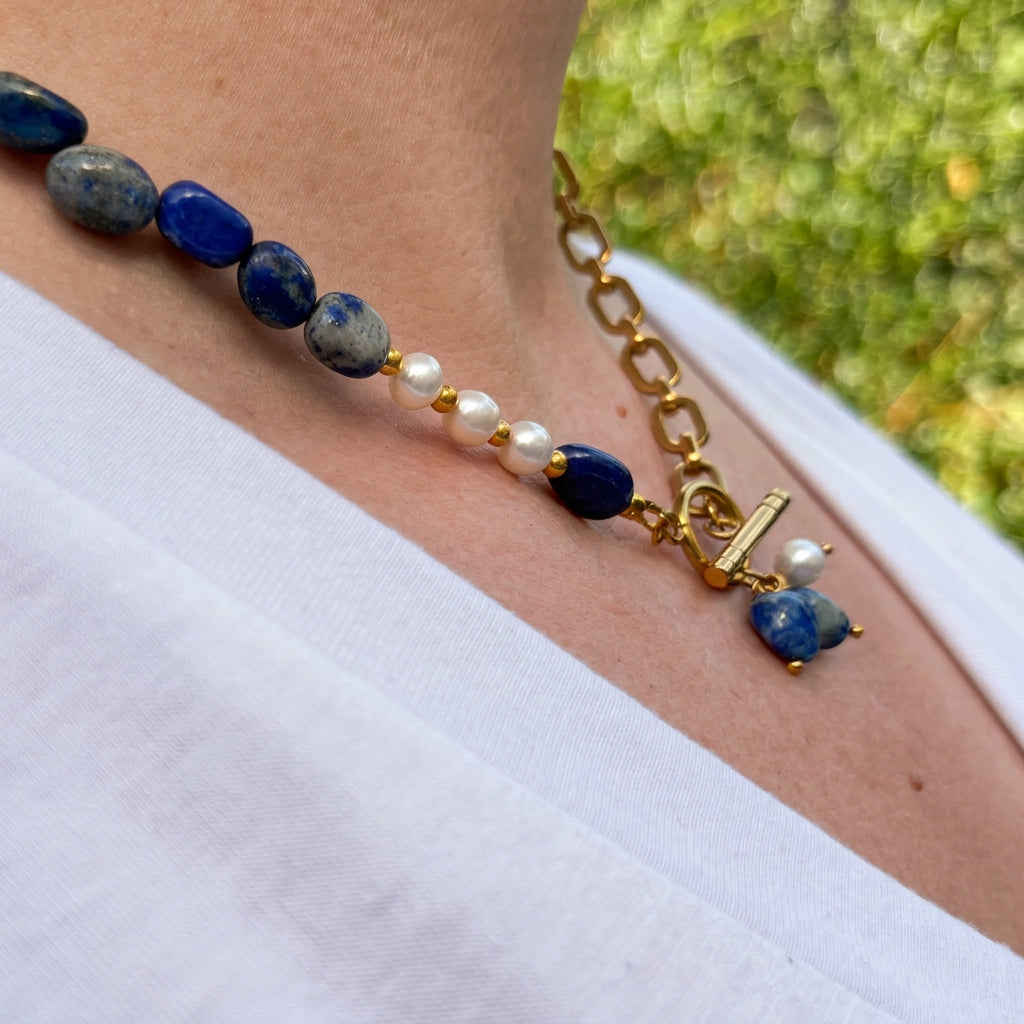 Side-angle view of a blue lapis lazuli and white baroque pearl necklace with a gold-plated paperclip chain, front toggle clasp and lapis-and-pearl pendant cluster, worn with a white V-neck top outdoors.