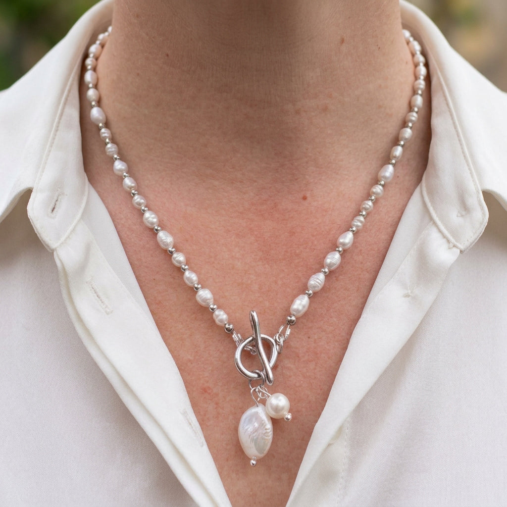 White rice pearl necklace with silver-coloured spacers, a fastened front toggle clasp and dangling coin and baroque pearls, worn with an open-neck white shirt.