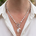 White rice pearl necklace with silver-coloured spacers, a fastened front toggle clasp and dangling coin and baroque pearls, worn with an open-neck white shirt.