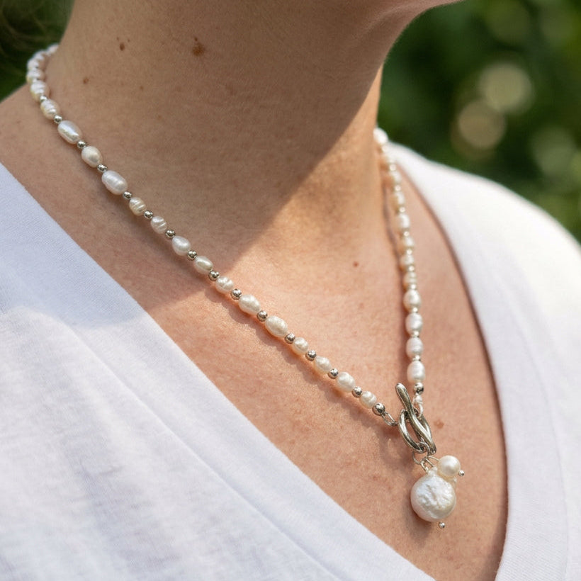 Side-angle view of a white rice pearl necklace with silver-coloured spacer beads, a front toggle clasp and dangling coin and baroque pearl cluster, worn with a white V-neck top outdoors.