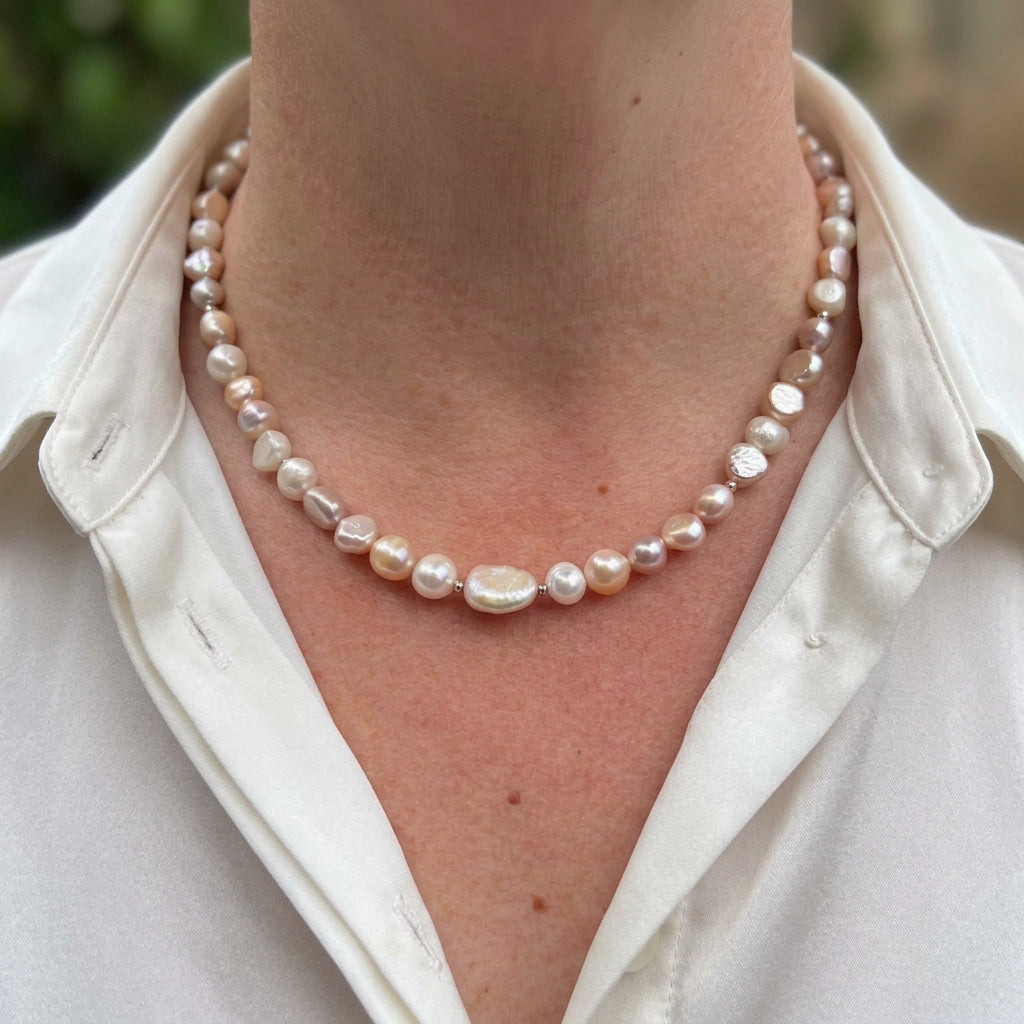 Cream, peach and pale lavender baroque pearl necklace with small sterling silver spacers and a larger flat pearl at the centre, worn at the collarbone with an open-neck white shirt.⠋