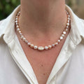 Cream, peach and pale lavender baroque pearl necklace with small sterling silver spacers and a larger flat pearl at the centre, worn at the collarbone with an open-neck white shirt.⠋