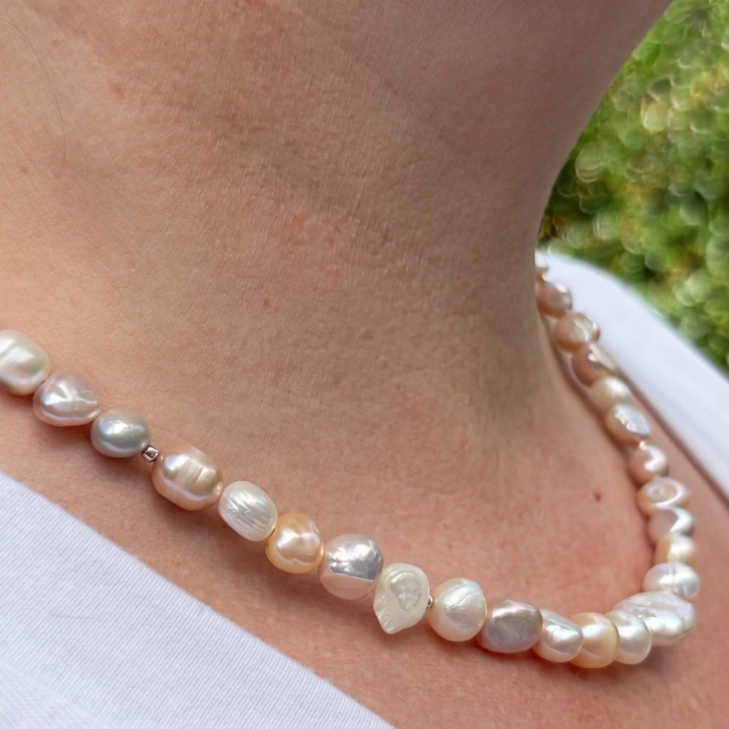 Side view of a multicoloured baroque pearl necklace worn with a white top, showing irregular cream, peach, pale gold and lavender pearls with small sterling silver spacers and an iridescent lustre.
