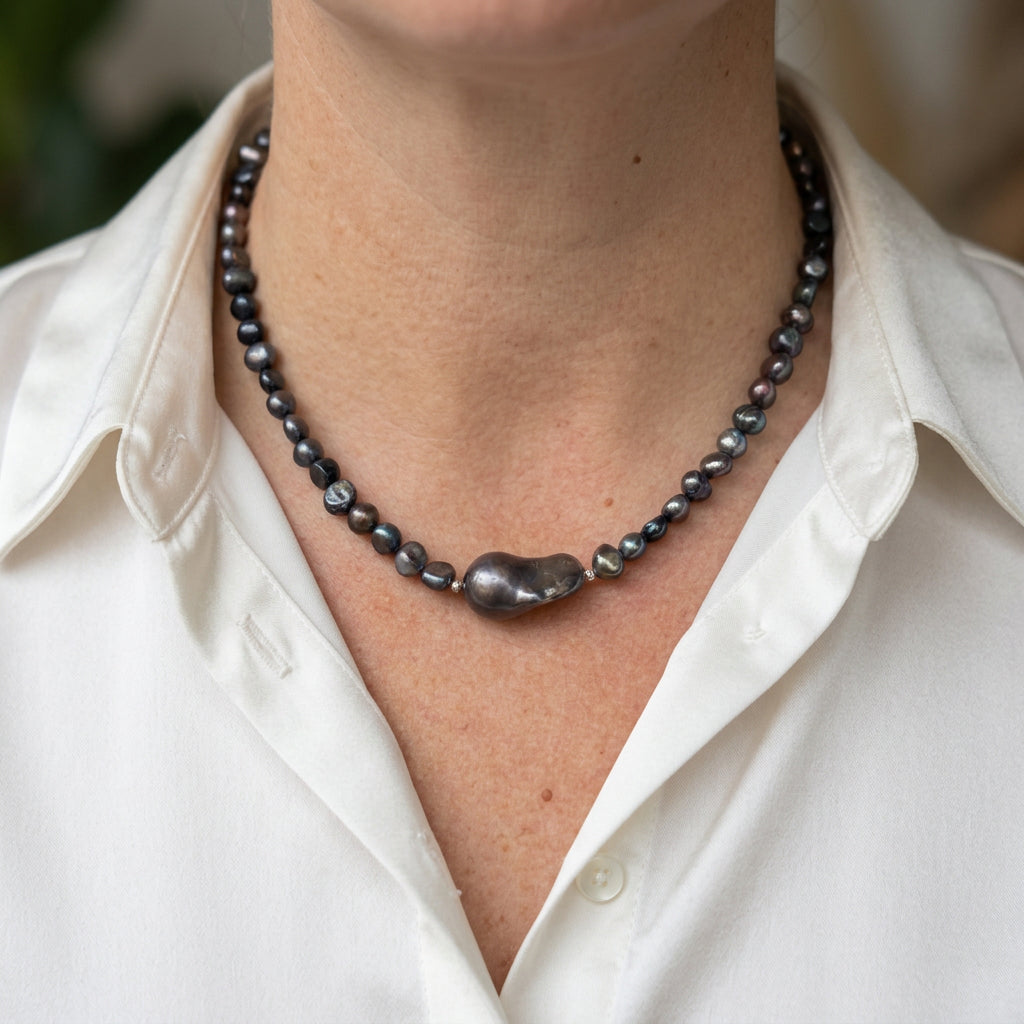 Dark baroque pearl and hematite necklace with a large irregular dark keshi pearl at the centre, worn with an open-neck white shirt, showing silvery iridescent highlights.
