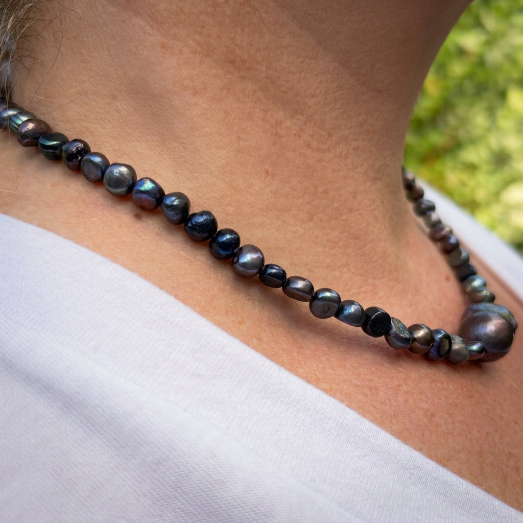 Side view of a dark blue baroque pearl and hematite necklace worn with a white V-neck top, showing irregular pearls with blue, green and purple reflections and a large dark keshi pearl centrepiece.