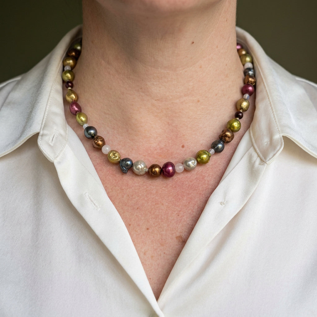 Multicoloured baroque pearl necklace in burgundy, bronze, lime-gold and silver-grey tones, interspersed with small pale gemstone beads, worn with an open-neck white shirt.