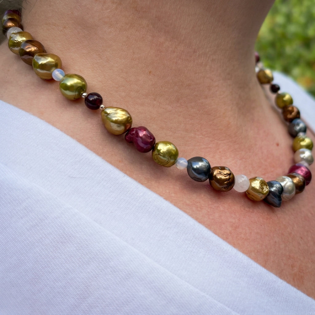 Side view of a multicoloured baroque pearl necklace worn with a white V-neck top, showing lime-gold, bronze, burgundy and silver-grey pearls with pale translucent gemstone accents and a dark red garnet bead.⠋