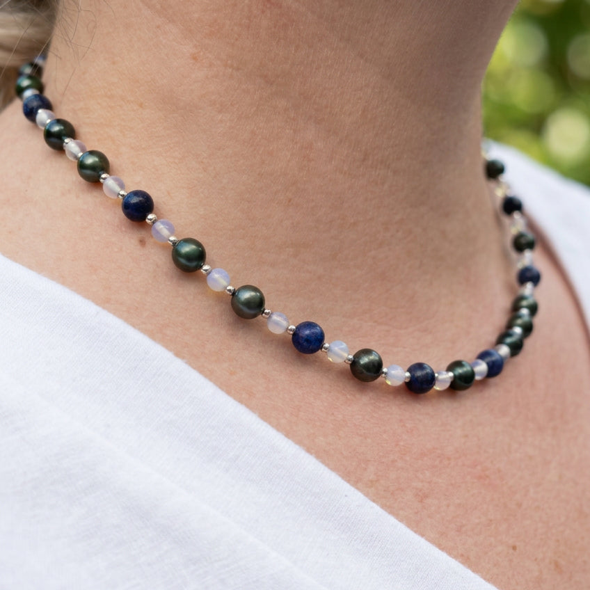 Side view of a peacock pearl, lapis lazuli and moonstone necklace worn with a white V-neck top, showing green-teal pearl lustre, deep blue stones, pale translucent beads and small sterling silver spacers.