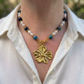 Mottled blue jasper beads and pairs of white baroque pearls with gold-coloured hematite spacers, a fastened front toggle and large gold-plated flower pendant, worn with an open-neck white shirt.
