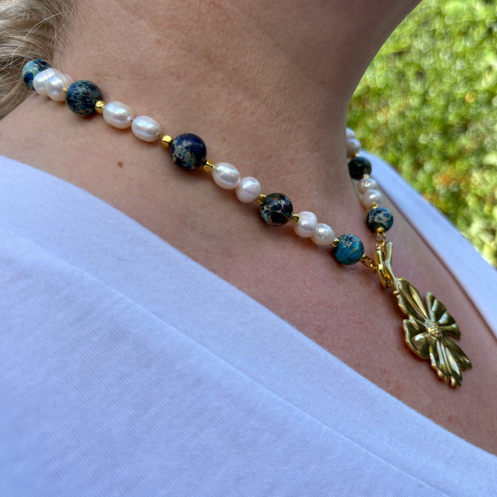 Side view of a blue jasper and white baroque pearl necklace worn with a white V-neck top, showing mottled blue beads, gold-coloured hematite spacers and a large gold-plated flower pendant beneath the front toggle clasp.⠋