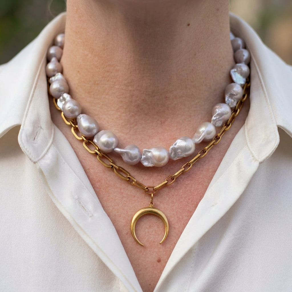 Double-layer necklace with silvery-white keshi pearls above a gold-plated paperclip chain and downward-pointing crescent moon pendant, worn with an open-neck white shirt.