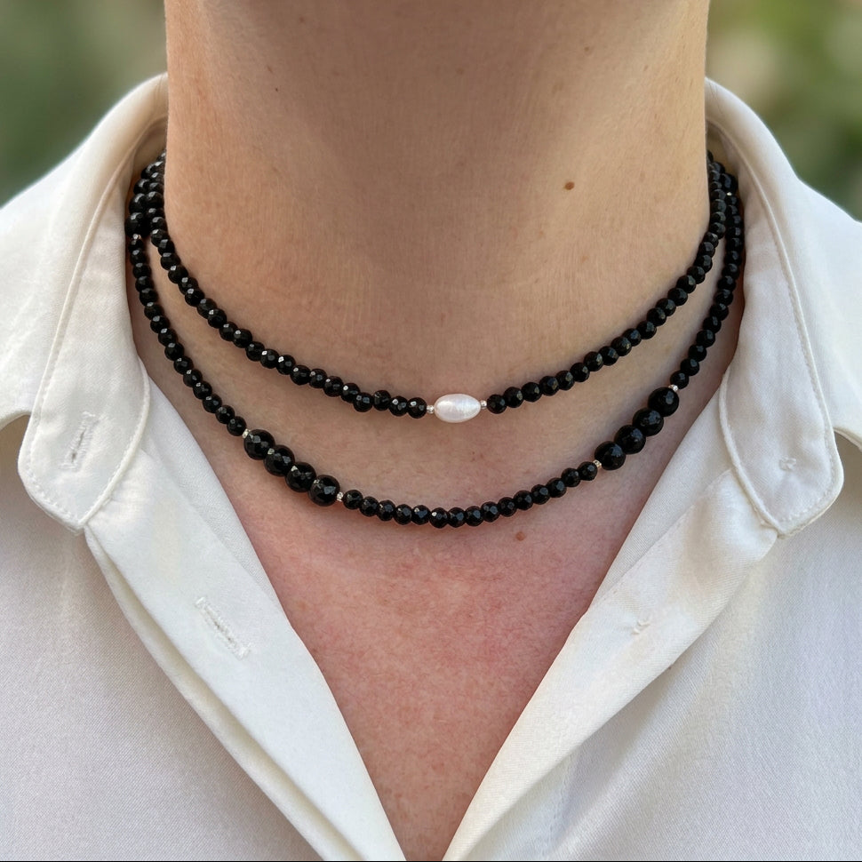 Two layered faceted black onyx necklaces worn with an open-neck white shirt, featuring a single white baroque pearl on the shorter strand and mixed-size beads on the longer strand.