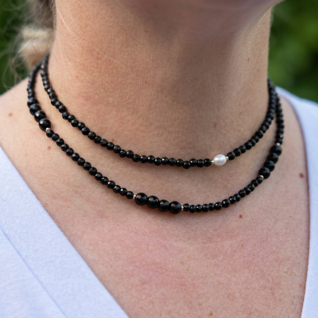 Angled close-up of two layered black onyx necklaces worn with a white top, showing a white baroque pearl on the shorter strand and groups of larger faceted beads on the longer strand.