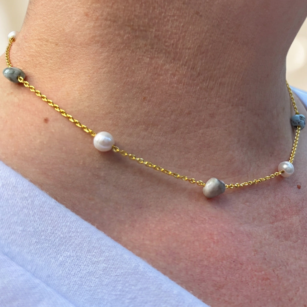 Angled close-up of a pearl and larimar station necklace worn with a white top, showing alternating white baroque pearls and blue-grey stones along a gold-filled chain.⠋