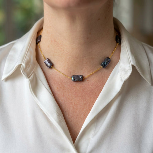 Dark blue rectangular baroque pearls spaced along a delicate gold-filled chain, worn with an open-neck white shirt, showing their textured surfaces and blue-green iridescent lustre.⠋